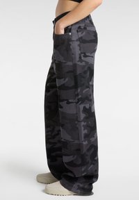 Camo pants in shades of gray and black, made of fabric with a smooth texture; relaxed fit with side pockets, worn with light-colored footwear.