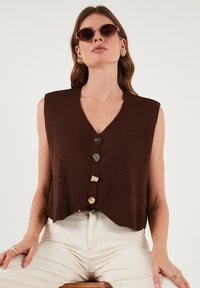 Brązowa krótka kamizelka z dekoltem w kształcie litery V, trzema złotymi guzikami i dwiema przednimi kieszeniami. Gładka tekstura, bezrękawnik, minimalistyczny styl.