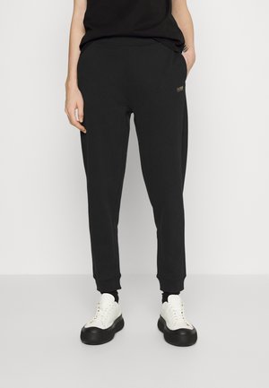 Person wearing black tapered sweatpants with elastic cuffs, black top, and white sneakers with black soles, standing against a plain background.