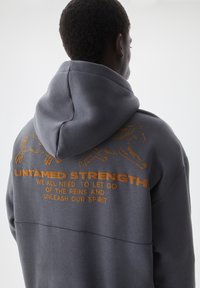 Grijze hoodie met een capuchon met trekkoord, met oranje tekst en paardenafbeeldingen op de achterkant. De stof lijkt zacht en gestructureerd.