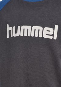 Grå sweatshirt med blå detaljer som har en stor vit "hummel"-logotyp. Materialet verkar mjukt med en slät textur. Enkel design med rundad halsringning.