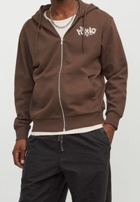 Felpa con zip marrone realizzata in tessuto morbido, dotata di tasca frontale, cappuccio con coulisse e una grafica bianca sulla parte sinistra del petto.