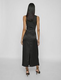 Abito in satin nero con top aderente e gonna svasata; presenta uno spacco centrale nella parte posteriore ed è abbinato a tacchi a spillo neri.