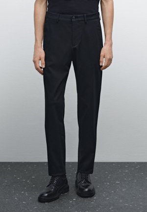 Black tailored pants made of a smooth fabric, featuring a straight fit, belt loops, and front pockets, paired with black lace-up shoes.