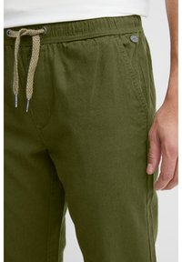 Mies, jolla on päällään oliivinvihreät housut, joissa on kiristysnyöri vyötäröllä ja sivutaskut, käsi rentona reidellä.