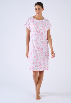 Frau steht barfuß und trägt ein weißes knielanges Kleid mit pinken und hellblauen Blattmustern und kurzen Ärmeln.