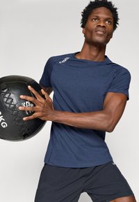 Jūras spēku sporta krekls ar īsām piedurknēm un šuvju detaļām, turēts ar melnu medicīnas bumbu ar teksturētu virsmu un logo.