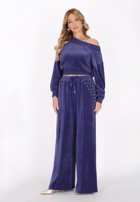 Lila velour träningsoverall med en avskuren off-shoulder topp med rhinestone detaljer och vida byxor med dragsko i midjan.