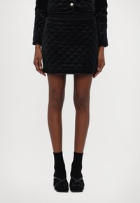 Mini gonna in velluto trapuntato nero con motivo a diamante, abbinata a calze nere fino alla caviglia e scarpe nere con tacco. Design semplice e aderente.