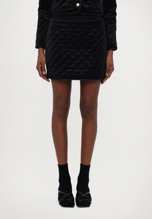 Mini-jupe en velours noir matelassé avec un motif en diamant, assortie à des chaussettes noires et des chaussures à talons noires. Design simple et ajusté.