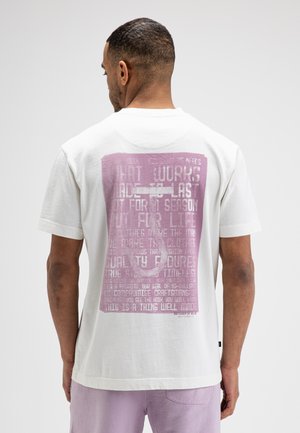 Mann trägt weißes T-Shirt mit großer pinker Grafik und Text auf dem Rücken, kombiniert mit hellvioletten Shorts, steht vor einfachem Hintergrund.