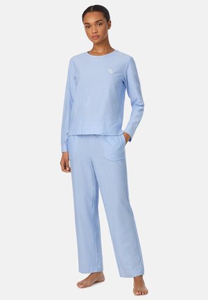 Mujer descalza, de pie, vistiendo un top de manga larga de rayas finas azul claro y blanco, y pantalones a juego con bolsillo.