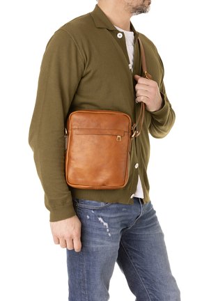 Borsa a tracolla in pelle color tan con una texture liscia, forma rettangolare, tasca frontale con zip e tracolla regolabile indossata su un cardigan color oliva.