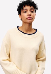Jongere met kort krullend haar, gekleed in een crèmekleurige trui met marineblauwe details, staande tegen een plain witte achtergrond.