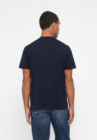 Man met zijn rug naar de camera, gekleed in een eenvoudig donkerblauw T-shirt met korte mouwen en blauwe jeans met lichte slijtage op de achterzakken.