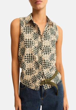 Blusa beige senza maniche con bottoni e motivi geometrici circolari neri, indossata con jeans blu e cintura intrecciata oliva.