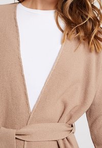 Beige stickad cardigan med V-ringning, texturerat tyg och knyts vid midjan. Det inre lagret är en vit skjorta, delvis synlig.