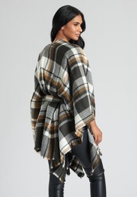 Rutigt poncho i svart, vitt och bruna nyanser; mjukt material med fransade kanter, öppen framside-design och lösa ärmar. Lämplig för lager-på-lager.