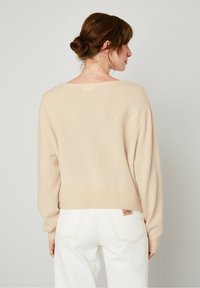 Pull beige clair à coupe décontractée, manches longues et encolure large. Associe à un pantalon en jean blanc avec une étiquette rouge sur la poche arrière.