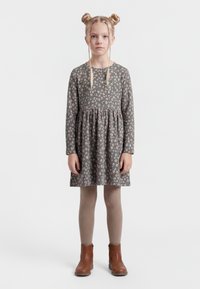 Ragazza con doppie trecce a chignon bionde, che indossa un vestito grigio a maniche lunghe con una piccola stampa floreale, collant beige e stivaletti marroni, in piedi in posizione eretta.