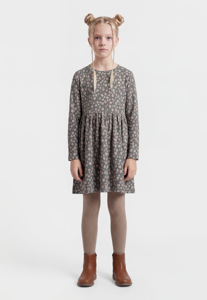 Ragazza con doppie trecce a chignon bionde, che indossa un vestito grigio a maniche lunghe con una piccola stampa floreale, collant beige e stivaletti marroni, in piedi in posizione eretta.