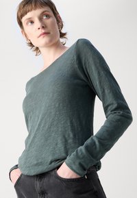 Ilgas rankovės marškinėliai prislopinto žalio atspalvio, pagaminti iš tekstūruoto audinio, su plačiu kaklo iškirpimu ir laisvu siluetu. Sujungti su tamsiomis džinsomis.