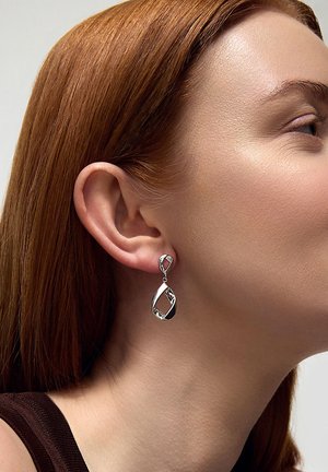 Srebrne kolczyki w kształcie kropli z misternym wycinanym wzorem, z akcentami wiszącymi, prezentowane na uchu modelki.
