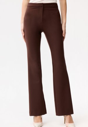 Kobieta ubrana w brązowe, rozszerzane spodnie z wysokim stanem, zestawione z białymi szpilkami w szpic, na jednolitym tle.