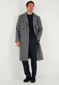 Grauer Wollmischung-Mantel mit doppelreihiger Vorderseite, Reverskragen und Seitentaschen, getragen über einem marineblauen Pullover und dunkler Hose.