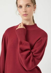 Czerwony sweter z wysokim dekoltem, z prążkowanymi mankietami i teksturowanym wykończeniem. Posiada bufiaste rękawy oraz luźny fason. Gładka tkanina.