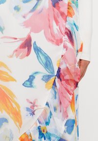 Tissu à motif floral en blanc avec de grandes fleurs roses, bleues et jaunes. Présente une texture superposée et transparente avec des bords froncés.