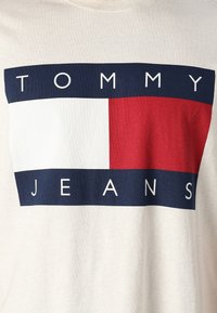 Beige t-särk, millel on julge graafiline disain, millel on sõnad "TOMMY JEANS" tume sinistes ja punastes plokkides ning eest valge ristkülikuline ala.