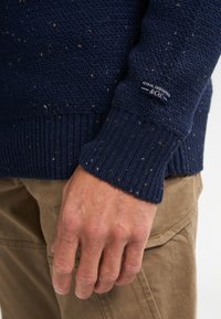 Pull en mailles bleu marine avec un motif texturé, des poignets côtelés et une étiquette de logo. Le tissu présente de subtiles touches de couleurs variées.