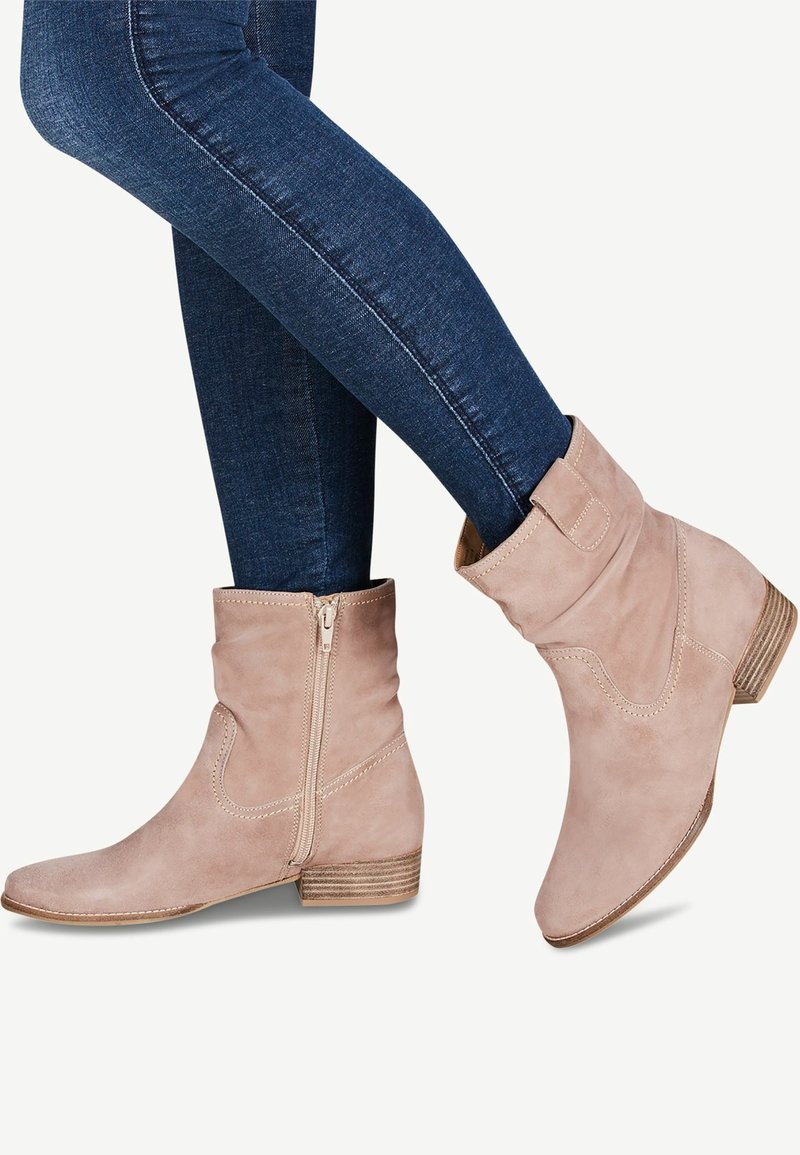 Bottines en daim beige avec fermeture éclair latérale, portées avec un jean skinny bleu foncé, montrant un petit talon et des détails cousus.