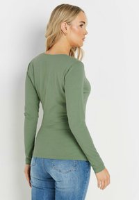 Langärmliges, olivgrünes Oberteil mit rundem Halsausschnitt und glatter Textur, kombiniert mit blauen Denim-Jeans, die feine Nähte aufweisen.