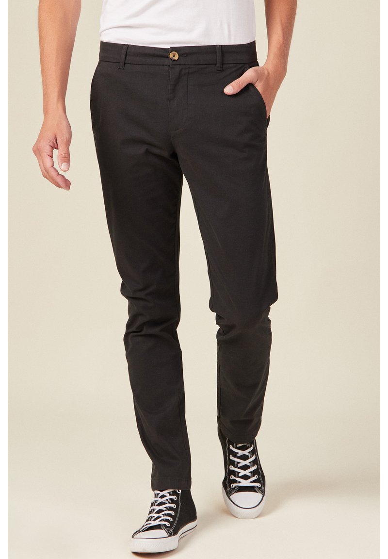 Schwarze Baumwollhosen mit slim fit, ausgestattet mit einem Knopfverschluss, seitlichen Taschen und kontrastierenden Nähten. Kombiniert mit schwarzen Sneakers.