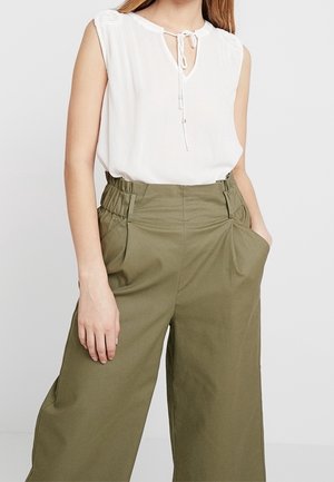 Donna con camicetta bianca senza maniche con dettaglio a fiocco e pantaloni a gamba larga, vita alta, verde oliva con tasche.