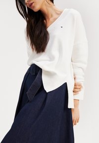 Pull en tricot blanc avec un col en V, doté de bords côtelés et d'entailles sur les côtés, associé à une jupe en denim foncé avec une taille ceinturée.