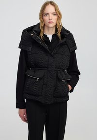 Czarna kamizelka puchowa z teksturowaną, falistą powierzchnią, zapinanymi kieszeniami i kapturem. Posiada epaulety zapinane na zatrzaskach na ramionach.