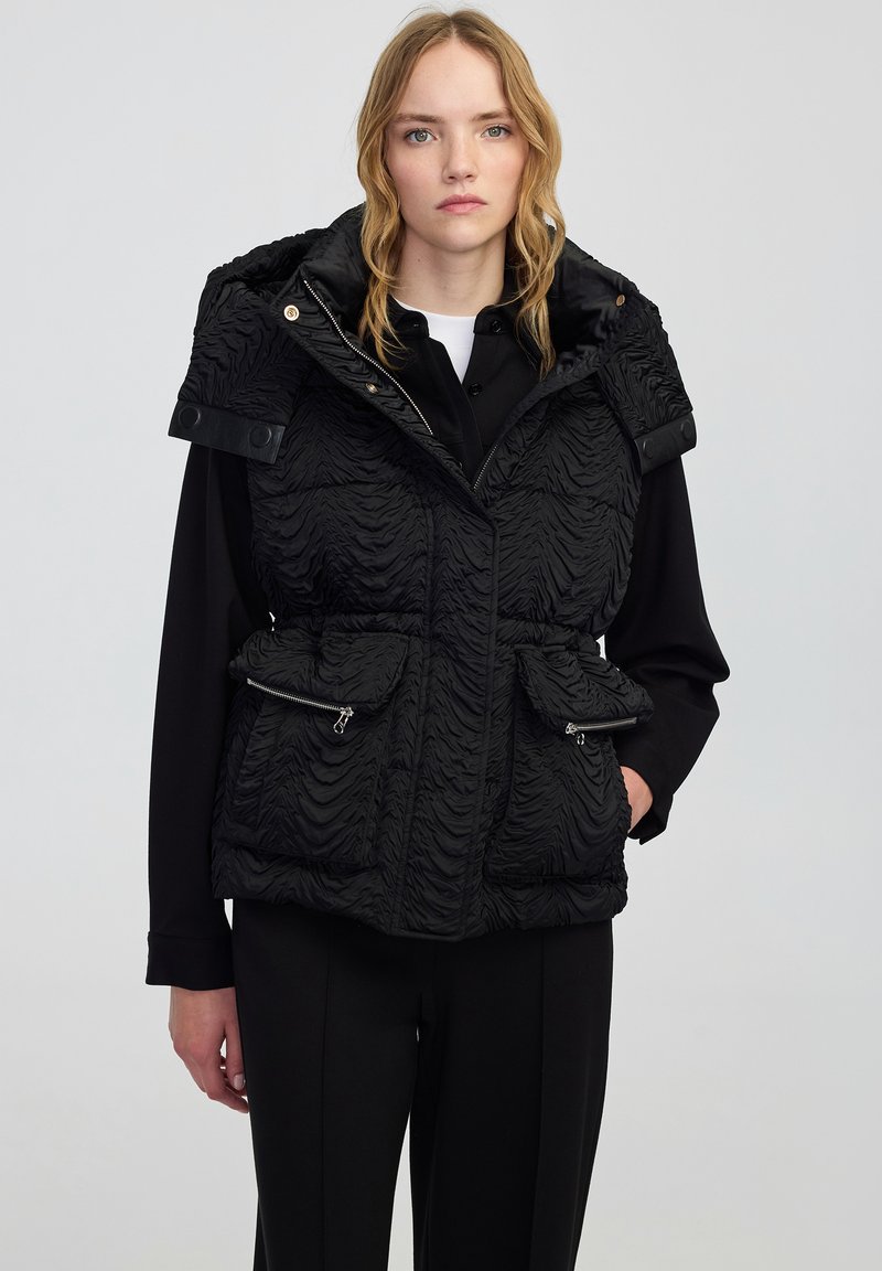 Czarna kamizelka puchowa z teksturowaną, falistą powierzchnią, zapinanymi kieszeniami i kapturem. Posiada epaulety zapinane na zatrzaskach na ramionach.
