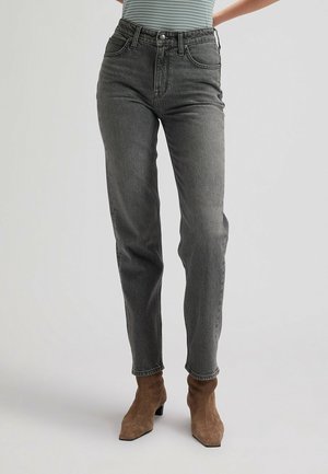 Osoba nosząca szare jeansy z wysokim stanem i brązowe zamszowe botki, stojąca na tle jednolitego jasnego tła.