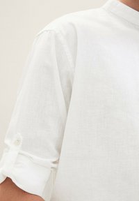 Weißes Leinenhemd mit Stehkragen, kurzen Ärmeln und hochgekrempelten Bündchen. Enthält einen weichen, strukturierten Stoff mit einem subtilen Webmuster.