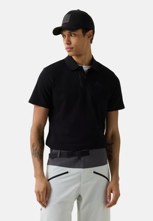 Mann mit schwarzer Kappe und Poloshirt mit grauer und weißer Hose, stehend vor hellem, einfarbigem Hintergrund, blickt zur Seite.