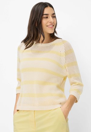 Frau lächelt, trägt einen hellgelben, gestreiften, locker gestrickten Pullover mit Dreiviertelärmeln und passende gelbe Hose, Hände in den Taschen.