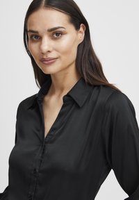 Schwarzes Hemd mit Knopfleiste aus glattem Stoff und klassischem Kragen. Verfügt über eine verdeckte Knopfleiste und lange Ärmel, entworfen für eine figurbetonte Silhouette.