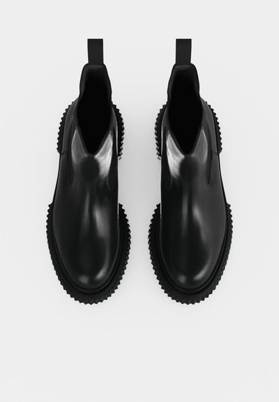 Paire de bottines en cuir noir brillant à enfiler avec des semelles épaisses rainurées et des languettes à l’arrière, vues de dessus sur fond blanc.