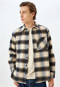 Flannel skjorte i sort, gul og grå med et ternet mønster. Har knaplukning foran, krave og en enkelt brystlomme.