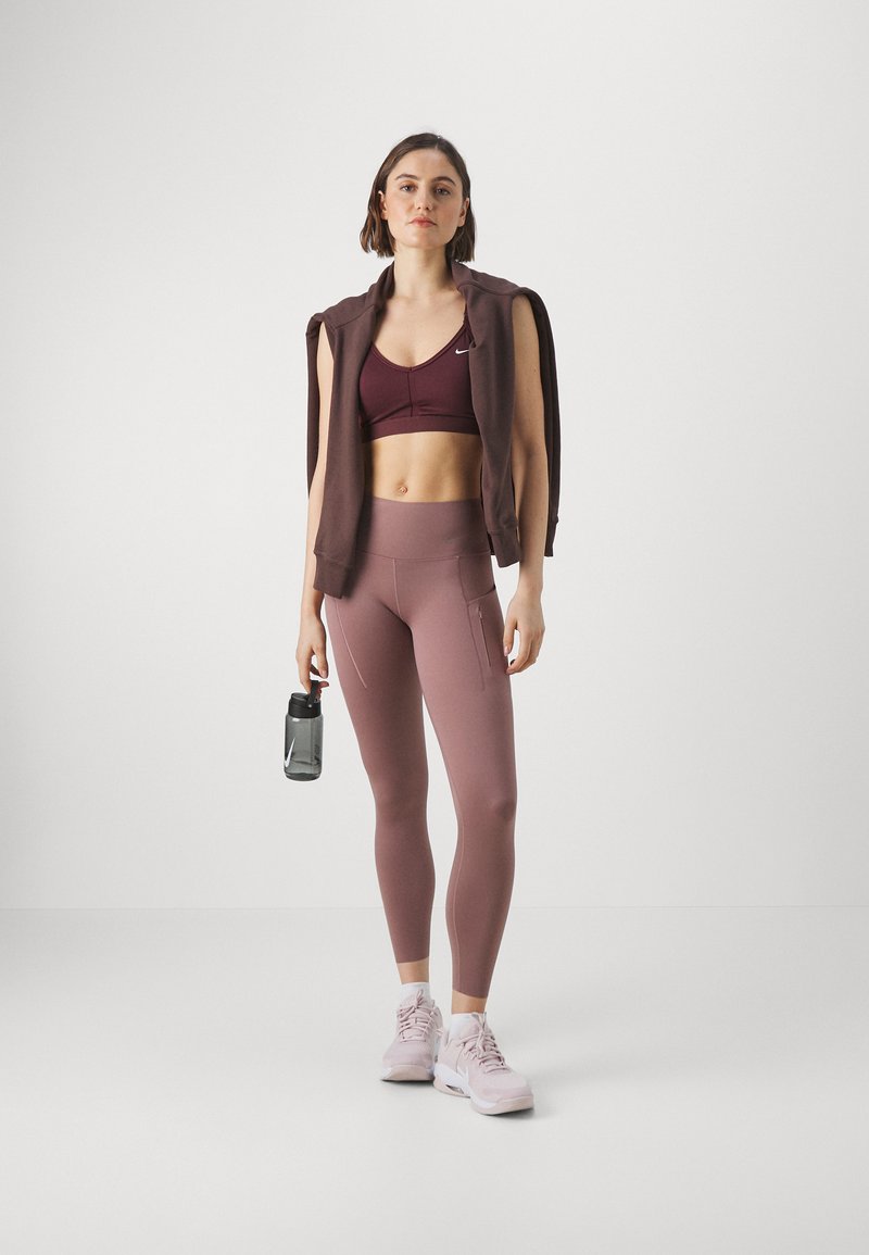 Conjunto deportivo que incluye un sujetador deportivo burdeos, leggings rosa empolvado con bolsillos laterales y una sudadera marrón caída sobre los hombros. Sosteniendo una botella de agua.