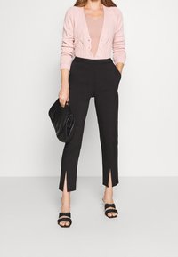 Schwarze maßgeschneiderte Hose mit seitlichen Schlitzen am Saum, strukturierte rosa Strickjacke und schwarze Clutch, kombiniert mit schwarzen Sandalen mit Riemchen.