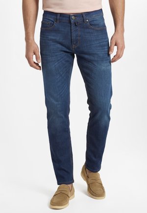 Mann trägt eng anliegende blaue Jeans und braune Loafers, steht vor weißem Hintergrund mit entspannten Händen seitlich.